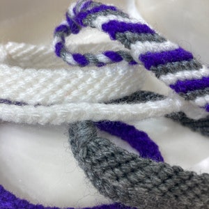 May include: Three braided cords in white, gray, and purple. The cords are woven together in a simple pattern.