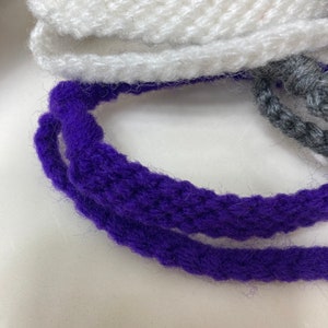 May include: Close-up of three strands of yarn, one white, one purple, and one gray. The yarn is soft and fluffy, and the colors are vibrant.