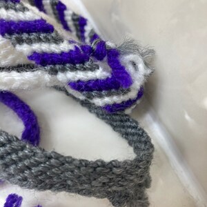 May include: A close-up of a gray, white, and purple woven bracelet. The bracelet is made of yarn and has a simple, striped pattern.