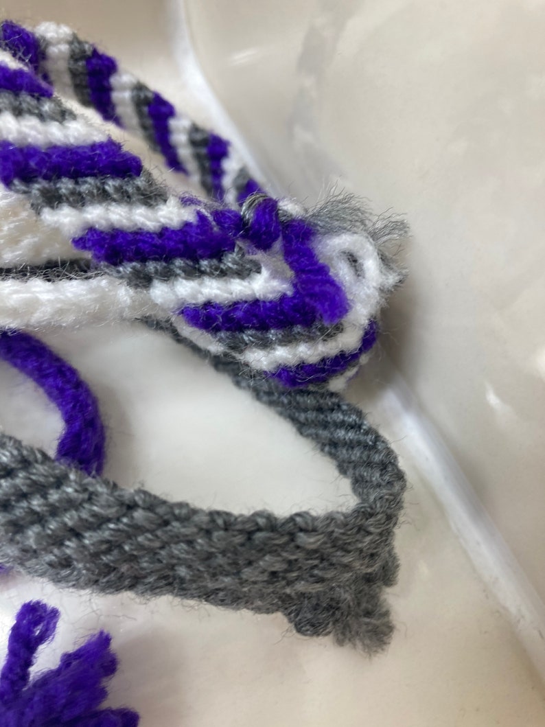 May include: A close-up of a gray, white, and purple woven bracelet. The bracelet is made of yarn and has a simple, striped pattern.