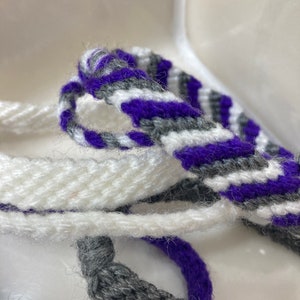 May include: A close-up of three woven friendship bracelets in white, gray, and purple. The bracelets are made with a simple, striped pattern.