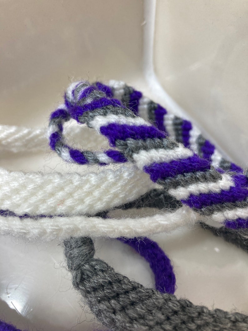 May include: A close-up of three woven friendship bracelets in white, gray, and purple. The bracelets are made with a simple, striped pattern.