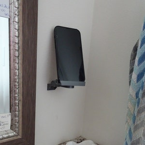 May include: A black phone is mounted on a white wall with a black phone holder. The phone is in portrait mode and the screen is black. The phone holder is attached to the wall with a screw.