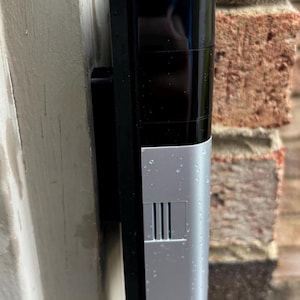 May include: A black and silver doorbell camera mounted on a brick wall. The camera has a rectangular shape and a small speaker grille.