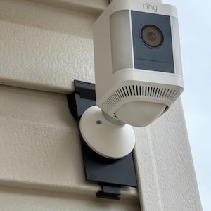May include: A white Ring security camera with a black mounting bracket. The camera is mounted on a white siding wall. The camera has a black lens and the word "ring" is printed on the top of the camera.
