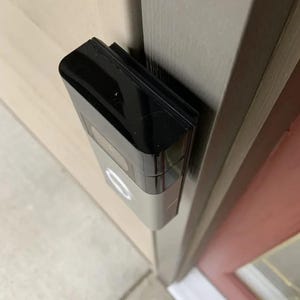 May include: A silver and black smart doorbell is mounted on a light-colored door frame. The doorbell has a rectangular shape with a black top and a silver body. The doorbell is installed on a light-colored door frame.