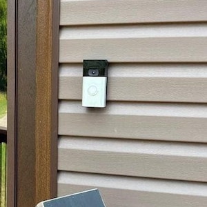 Peut inclure: Une sonnette intelligente blanche et noire est fixée sur un mur de bardage beige. La sonnette possède une caméra et un bouton. Le bardage est rainuré horizontalement. Une garniture en bois est sur le côté gauche de l'image.