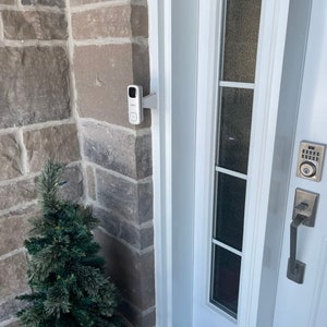 Può includere: Una porta bianca con una serratura a tastiera e una videocamera per il campanello montata sul muro accanto. Un piccolo albero sempreverde è in primo piano.