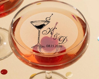 May include: Two clear glass coupe glasses filled with a reddish liquid and a single cherry. Each glass features a circular white topper printed with a black martini glass icon, the letters K & D, and the date 08.13.2026 in black text.