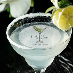 May include: A white margarita glass with a salted rim, filled with a clear liquid and a green olive garnish. A round sticker on the glass reads "Michelle's Birthday A TINI BIT OLDER".