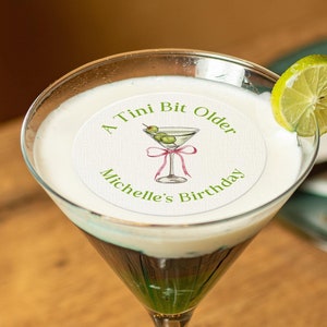 May include: A martini glass filled with a green and white layered drink topped with whipped cream. A round white sticker with green text reads "A Tini Bit Older Michelle's Birthday" and features a drawing of a martini glass with olives and a pink ribbon. A lime wedge is on the rim of the glass.