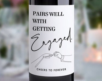 May include: A white wine bottle label with black text that reads "Pairs well with getting engaged."  The label also features a line drawing of two hands holding a ring.  The bottom of the label reads "Cheers to forever."