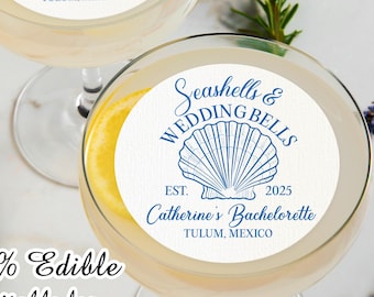 Puede incluir: Dos copas de cóctel transparentes con una rodaja de limón y un adorno comestible blanco. El adorno tiene un diseño de concha azul y el texto "Seashells & Wedding Bells" y "Catherine's Bachelorette Tulum, Mexico".