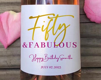 May include: A wine bottle with a white label featuring the words "Fifty & Fabulous" in gold and pink script. The label also includes "Happy Birthday Samantha" and the date July 07, 2023. A pink rose and petals are next to the bottle.