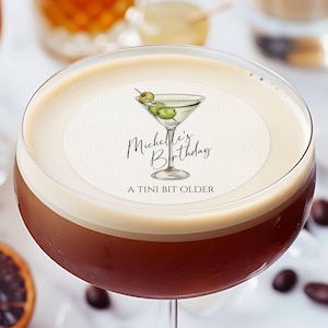 May include: A round edible image of a martini glass with olives, with the text "Michelle's Birthday A Tini Bit Older" printed on it. The image is on top of a brown drink in a martini glass. 100% Edible. Suitable for cupcake topper too.
