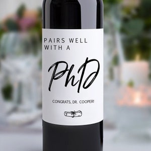 May include: A white wine bottle label with black text that reads "Pairs well with a PhD. Congrats, Dr. Cooper!" with a small illustration of a diploma scroll.