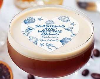 Op de afbeelding: Een cocktail in een glas met een eetbare topper in de vorm van een hart. De topper toont blauwe illustraties van schelpen en zeesterren met de tekst "SEASHELLS AND WEDDING BELLS Catherine's Bachelorette." De tekst "100% Edible Suitable for cupcake topper too" is ook aanwezig.
