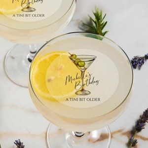 May include: Two martini glasses with lemon slices and cocktail napkins. The napkins have a hand-drawn illustration of a martini glass with olives and the text "Michelle's Birthday A TINI BIT OLDER".