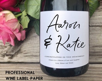 May include: A white wine bottle label with black calligraphy text that reads "Aaron & Katie" and the message "Congratulations on your engagement! Cheers to a lifetime of love and laughter. Love, Edison and Sheila".