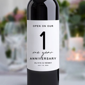 May include: A white wine bottle label with black text that reads "OPEN ON OUR 1 one year ANNIVERSARY OLIVIA & KENNY JULY 16, 2024".