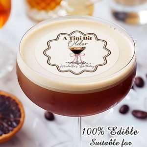 May include: A round edible image topper with the text "A Tini Bit Older" and a martini glass with coffee beans. The text "Michelle's Birthday" is below the martini glass. The topper is on top of a coffee drink in a glass. The text "100% Edible Suitable for cupcake topper too" is below the glass.