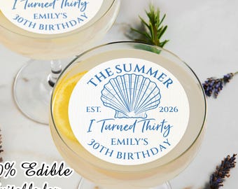 May include: Two clear cocktail glasses with a lemon slice and a white edible topper. The topper has a blue seashell design and text that reads "The Summer, I Turned Thirty, Emily's 30th Birthday". The text "100% Edible" is also visible.