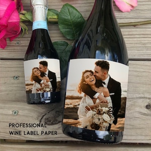Puede incluir: Dos botellas de vino con etiquetas personalizadas que muestran a una pareja de bodas. Las etiquetas est&aacute;n impresas en papel de etiqueta de vino profesional. Las botellas est&aacute;n sobre una superficie de madera con p&eacute;talos de rosa rosa y blanca.