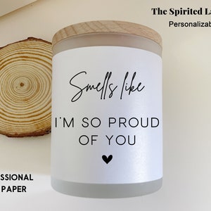 May include: White candle with a wooden lid and a label that says "Smells like I'm so proud of you" with a small black heart.