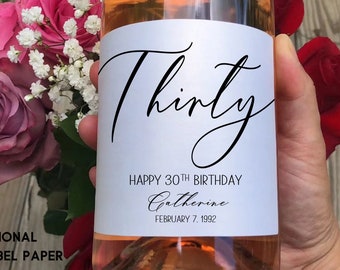 May include: A white wine bottle label with black calligraphy that reads "Thirty" and "Happy 30th Birthday Catherine February 7, 1992".