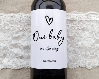 May include: A white wine bottle label with black text that reads "Our baby is on the way... DUE JUNE 2023". A black heart is drawn above the text.