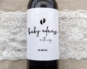 May include: A white wine bottle label with black text and a footprint symbol. The text reads "baby adams on the way. DUE JUNE 2022".