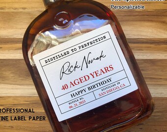Könnte beinhalten: Ein weißes Etikett mit schwarzem Text, der "Distilled to Perfection" und "40 Aged Years" lautet, mit einer handschriftlichen Unterschrift, die "Rick Novak" lautet. Das Etikett befindet sich auf einer braunen Glasflasche mit einer roten Flüssigkeit darin. Das Etikett enthält auch den Text "Happy Birthday", "Bottle No. 06.21.2023", "Distilled in San Diego, CA".