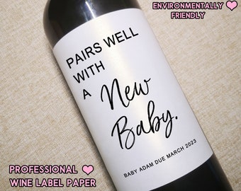 May include: A white wine bottle label with black text that reads "Pairs well with a New Baby. Baby Adam Due March 2023". The label also includes the text "The Spirited Labels Environmentally Friendly".