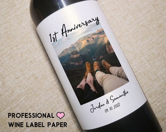 May include: A white wine bottle label with a photo of a couple sitting on a mountaintop with the text "1st Anniversary" above the photo and "Jordan & Samantha 09.10.2022" below the photo.