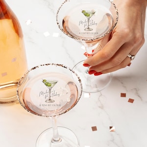 May include: Two martini glasses with gold rims and a personalized sticker that says "Michelle's Birthday A TINI BIT OLDER" with a drawing of a martini glass with olives.