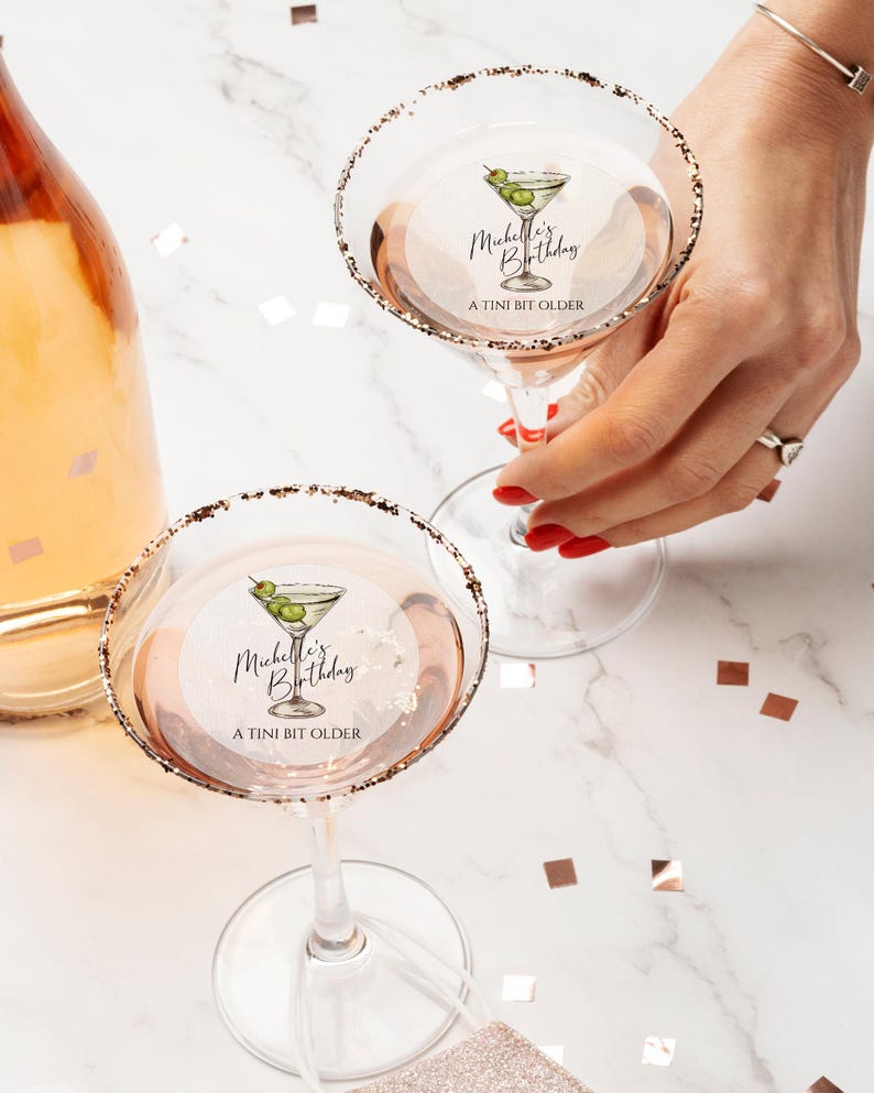 May include: Two martini glasses with gold rims and a personalized sticker that says "Michelle's Birthday A TINI BIT OLDER" with a drawing of a martini glass with olives.