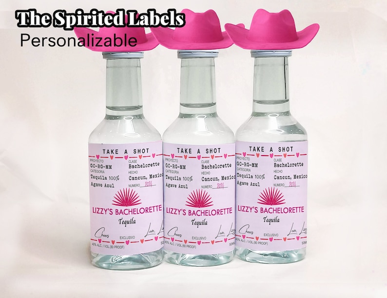 May include: Three clear glass bottles with pink and white labels. The labels have the text "Take a Shot" and "Lizzy's Bachelorette Tequila". Each bottle has a pink cowboy hat on top.