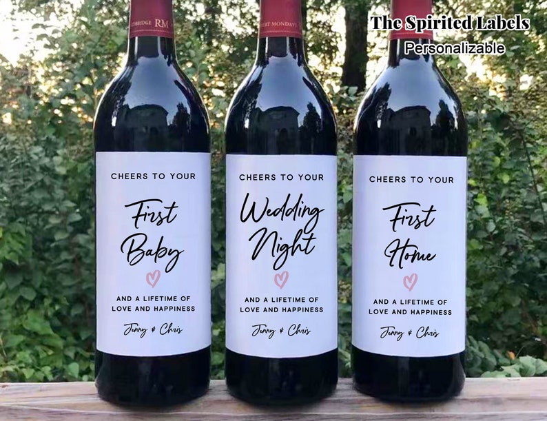 May include: Three wine bottles with personalized labels. The labels are white with black text and a pink heart. The text on the labels reads "Cheers to your First Baby", "Cheers to your Wedding Night", and "Cheers to your First Home".