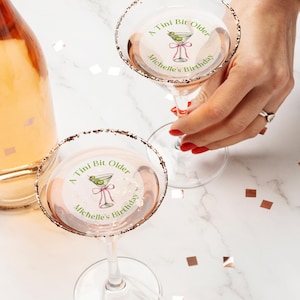 May include: Two clear glass martini glasses with gold rims. Each glass has a round sticker with a green martini glass illustration and the text "A Tini Bit Older Michelle's Birthday".