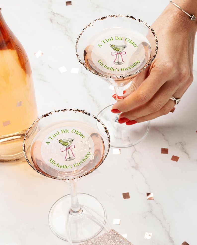 May include: Two clear glass martini glasses with gold rims. Each glass has a round sticker with a green martini glass illustration and the text "A Tini Bit Older Michelle's Birthday".