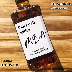 May include: A white wine bottle label with black text that reads "Pairs well with a MBA. Congrats David! We're So Proud Of You!" The label is on a brown glass bottle.