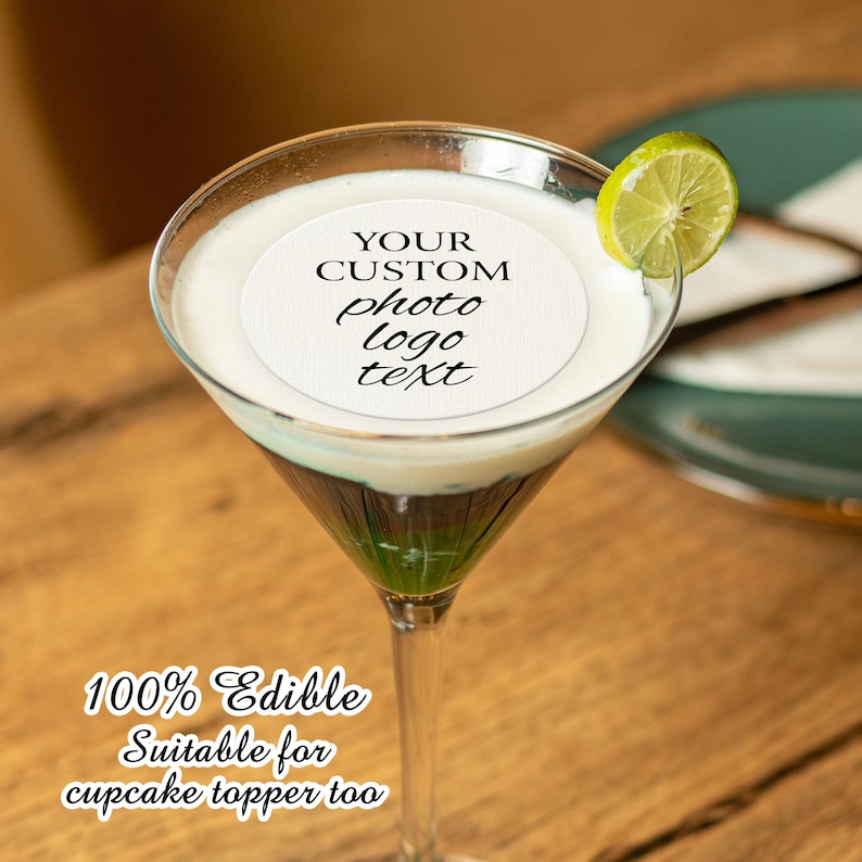 May include: A white round edible wafer with the text "YOUR CUSTOM photo logo text" on top of a green and white layered cocktail in a martini glass. The glass is on a wooden surface. 100% Edible. Suitable for cupcake topper too.
