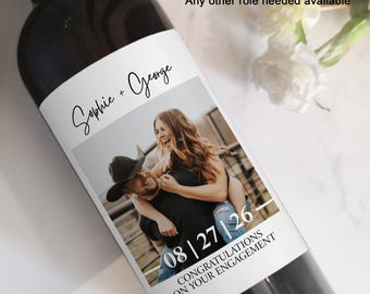 May include: A white wine bottle label featuring a photograph of a couple, the text "Sophie + George", and the date "08|27|26". The label also includes the text "Congratulations on your engagement".