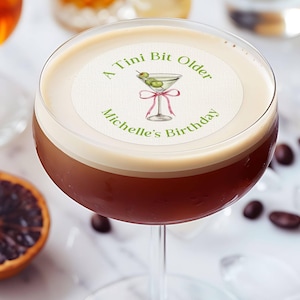 May include: A white round sticker with green text that reads "A Tini Bit Older Michelle's Birthday". The sticker has a green illustration of a martini glass with olives and a pink ribbon.
