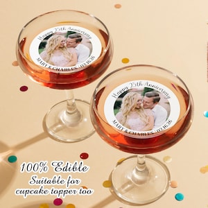 May include: Two clear glass champagne flutes with edible cupcake toppers. The toppers are round and feature a photo of a couple with the text "Happy 25th Anniversary Mary & Charles 02.16.26".