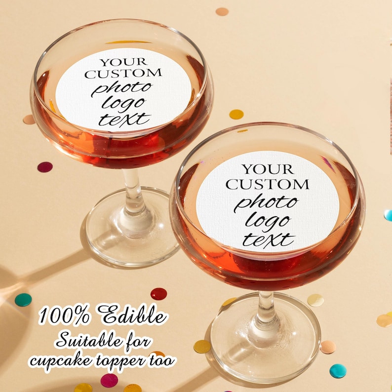 May include: Two clear glass champagne coupes with edible, round, white paper toppers. The toppers have black text that reads "YOUR CUSTOM photo logo text". The text is written in a cursive font. The glasses are filled with a red liquid. The background is a light beige with colorful confetti.