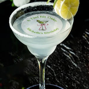 May include: A round, white, paper cocktail napkin with green text that reads "A Tini Bit Older Michelle's Birthday" and a green illustration of a martini glass with olives. The napkin is placed on top of a margarita glass filled with a clear liquid and a lime wedge.