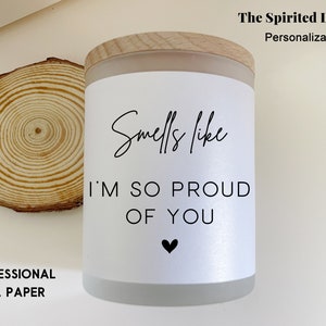 May include: White candle with a wood lid and a white label that says "Smells like I'm so proud of you" with a small black heart.