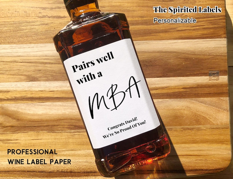 May include: A white label with black text on a brown glass bottle. The label reads "Pairs well with a MBA. Congrats David! We're So Proud Of You!" The Spirited Labels. Personalizable.