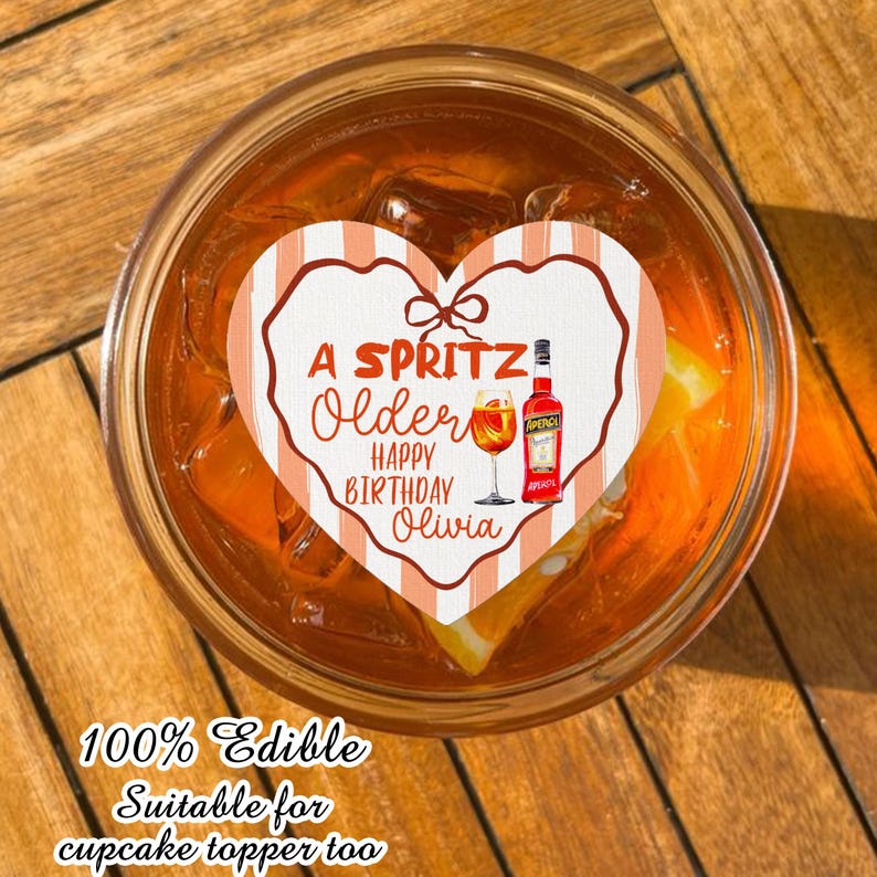 May include: A heart-shaped cupcake topper with the text "A Spritz, Older, Happy Birthday Olivia." The topper features a bow, a bottle, and a glass of Aperol. The drink is in a glass with ice and an orange slice.